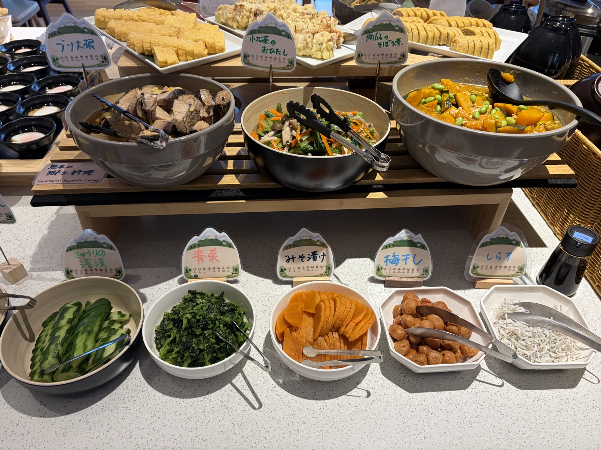たびのホテル阿蘇熊本空港の朝食ビュッフェ。ブリ大根、小松菜のおひたし、南瓜のそぼろ煮などの熊本郷土料理が、可愛い案内札とともにズラリと並んでいます。