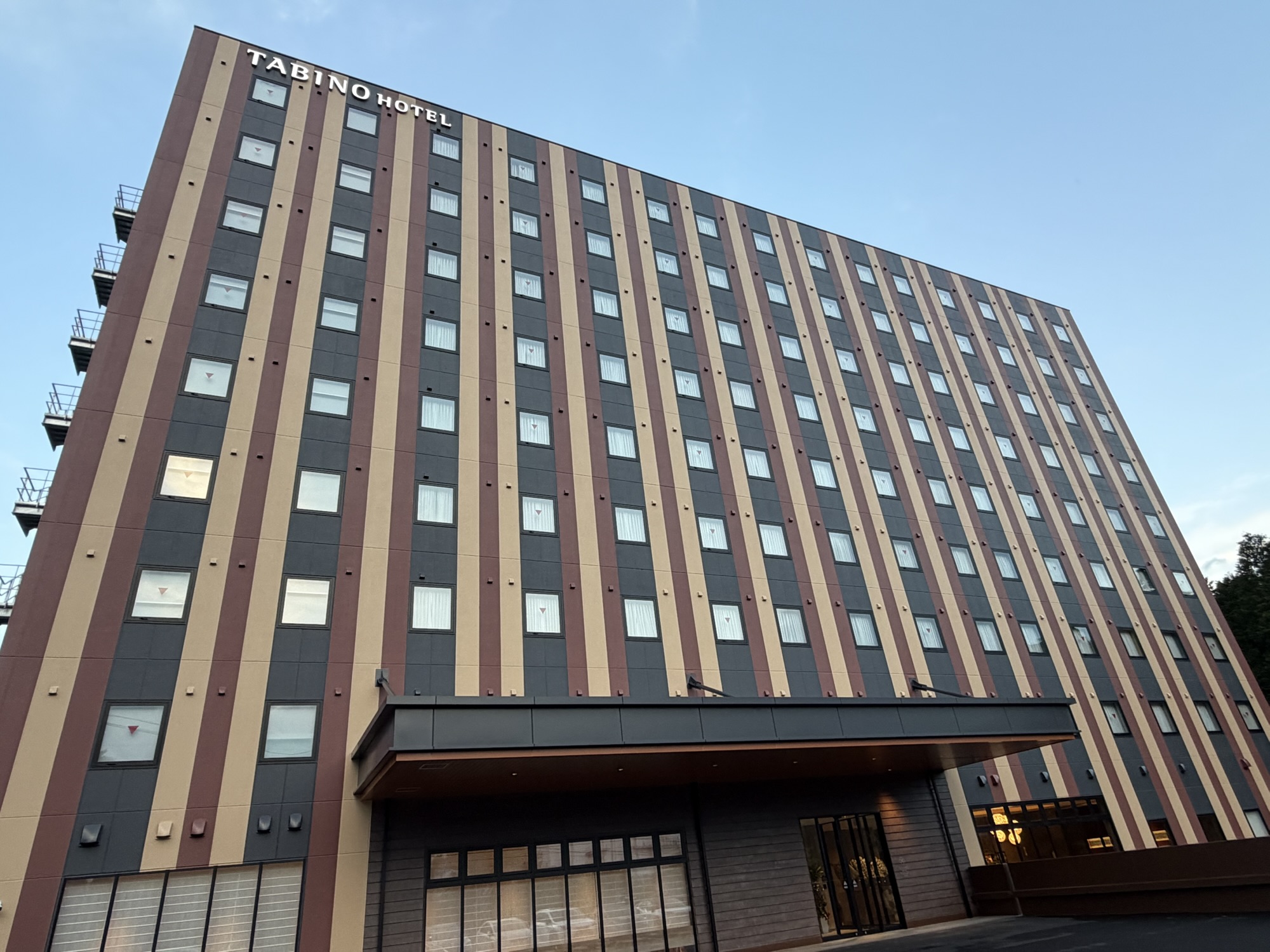2026年4月開業「たびのホテル阿蘇熊本空港」の外観。茶色とベージュの縦ストライプ柄が特徴的なモダンな建物で、屋上に「TABINO HOTEL」の白いロゴがあります。